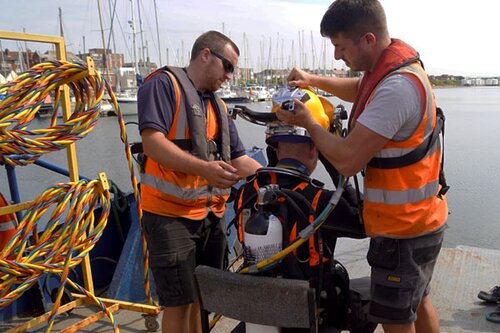 Two man in high vis jackets, helping a diver put on his kit and helmet.