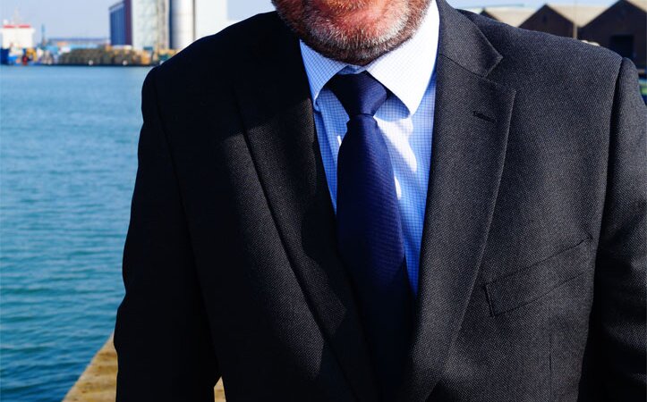 A white man in a dark suit, with a white shirt and dark navy tie, the port chimney is in the background.