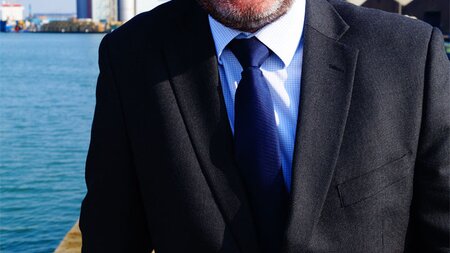 A white man in a dark suit, with a white shirt and dark navy tie, the port chimney is in the background.