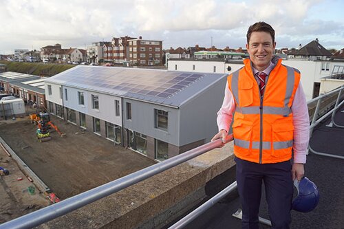 A man is stood on a flat roof balcony, overlooking a grey property.