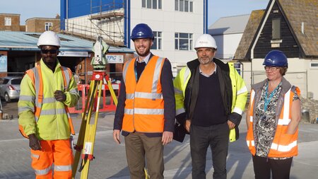 Four people standing in a row, one person is holding a level. They all wear high vis and hard hats.