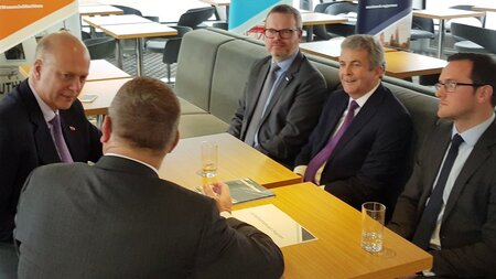 Five men in dark suits sit around a table talking to one another.