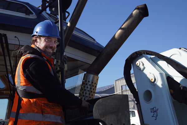 A marine colleague wearing an orange high vis jacket and a blue helmet. They are stood on the front of a ship.
