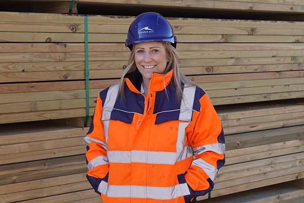 A women in an orange high vis jacket and blue helmet, stood in front a pile of timber.